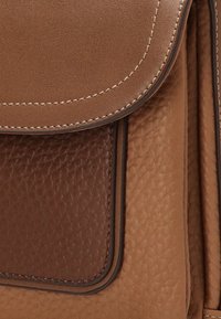 Bolso de mano de cuero marrón con una textura de grano fino, tapa suave y acentos oscuros en contraste. Los bordes cosidos resaltan los detalles del diseño.