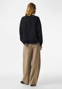 Femme debout, dos à l’objectif, portant un haut noir ample à manches longues, un pantalon beige à jambes larges et des chaussures noires sur un fond blanc uni.