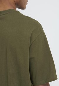 Tricou din bumbac verde măsliniu, cu mâneci scurte, guler rotund, textură vizibilă și detalii de cusătură pe umăr și corp.