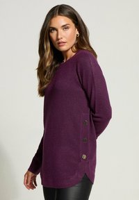 Lila Pullover mit rundem Ausschnitt, langen Ärmeln und drei dekorativen Knöpfen an der Seite. Hergestellt aus weichem, strukturiertem Stoff.