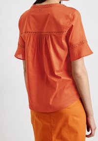 Blusa corta rossa-arancione con maniche a raglan arricciate sul retro, trim in pizzo sulle maniche e un bottone sul collo, realizzata in tessuto di cotone leggero.