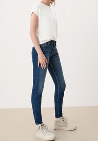 Úzké džíny z tmavě modrého denimu s jemným vyblednutím, doplněné o bílé tričko s krátkým rukávem a béžové vysoké tenisky.