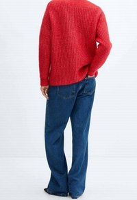 Personne se tenant de dos, portant un pull rouge texturé et un jean bleu ample avec une main dans la poche.