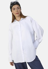 Camisa blanca de botón con un corte relajado, mangas largas y una textura suave, combinada con pantalones azul marino y un pañuelo negro.