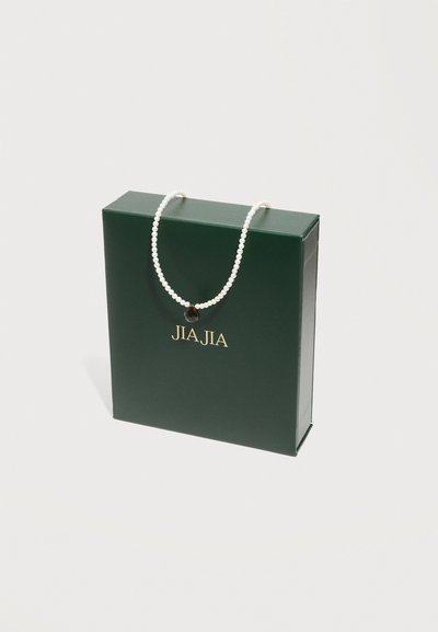 Κολιέ με μαργαριτάρια και σκουλαρίκι σε σχήμα σταγόνας με σκοτεινή απόχρωση, τοποθετημένο πάνω σε σκούρο πράσινο κουτί με την επιγραφή "JIA JIA" σε ανοιχτό φόντο.