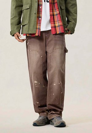 Uomo con pantaloni cargo marroni macchiati di vernice, sneaker grigie, giacca verde e sciarpa a quadri rossa sopra una camicia bianca.