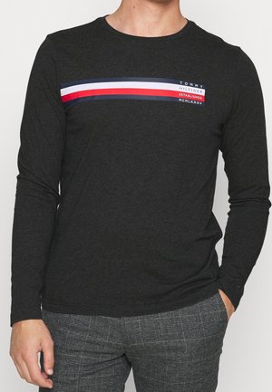 Mężczyzna ubrany w ciemną koszulę z długim rękawem z poziomymi paskami w kolorach granatowym, białym i czerwonym oraz nadrukiem Tommy Hilfiger na piersi, w połączeniu z szarą kratkowaną spodnią.