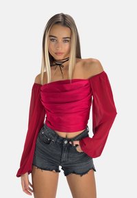 Haut rouge décolleté avec fronces, manches longues transparentes, associé à un short en denim noir déchiré. Tissu lisse avec une coupe ajustée.