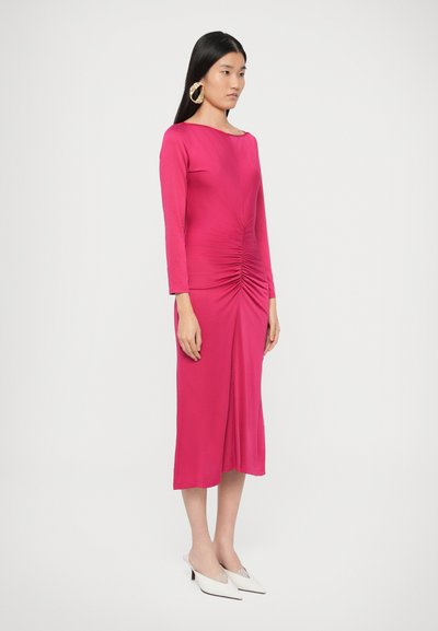 Rózsaszín hosszú ujjú ruha, elöl ráncolt részlettel és testhez simuló sziluettel. Fehér, hegyes orrú magassarkú cipővel viselve. Sima anyagtextúra.