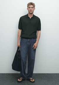 Mörkgrön, ribbade polo skjorta parat med löst sittande blå jeans. Bär en svart tote-väska; har på sig svarta flip-flops. Enkel design.