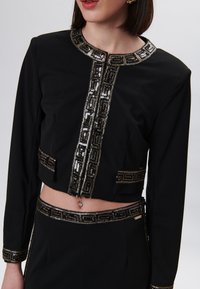 Vrouw in een zwart cropped jack en een rok met gouden en zwarte paillettenrand, met een navelpiercing en gouden oorbellen.