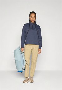 Granatowy polar z półzamek, beżowe spodnie, beżowe buty trekkingowe oraz jasnoniebieski plecak z regulowanymi paskami.