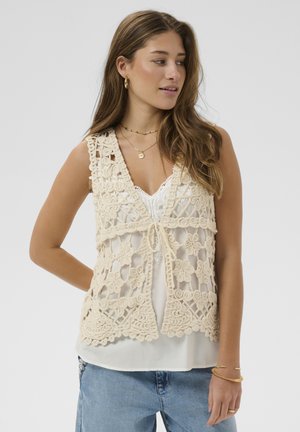 Młoda kobieta ubrana w kremową kamizelkę na szydełku na białej bluzce, niebieskie dżinsy, złote kolczyki koła oraz warstwowe naszyjniki, patrzy w lewo.