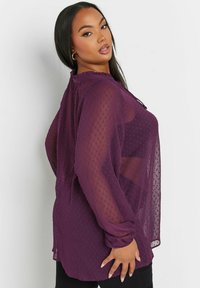 Blusa violacea trasparente con una texture a pois, colletto arricciato, maniche lunghe e dettagli con polsini arricciati, indossata sopra pantaloni neri.