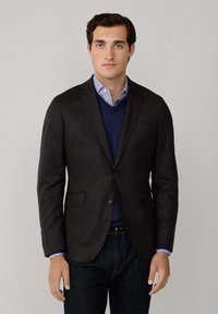 Blazer scuro realizzato in tessuto testurizzato, due tasche frontali, chiusura con un bottone, indossato sopra un maglione blu navy e una camicia azzurra, abbinato a jeans scuri.