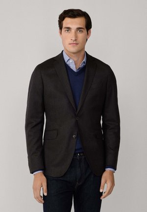 Blazer sombre en tissu texturé, avec deux poches avant, fermeture à un bouton, superposé sur un pull marine et une chemise bleu clair, associé à un jean foncé.