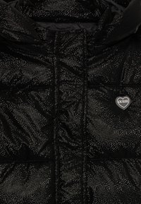 Veste en duvet noire avec des speckles réfléchissants, mettant en valeur un texture matelassée. Présente un logo en forme de cœur sur la poitrine.