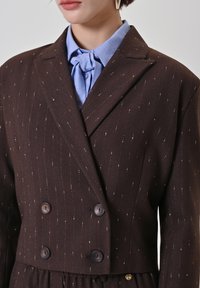 Brauner zweireihiger Blazer mit dünnen goldenen Nadelstreifen, ausgestattet mit einem Reverskragen und großen dunklen Knöpfen. Getragen über einer hellblauen Bluse.