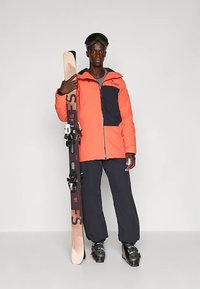 Skiklær med en oransje vanntett jakke med mørkeblå detaljer, kombinert med svarte skibukser og svarte skistøvler, som holder et par ski.