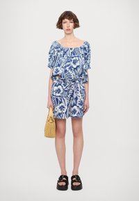 Blå og hvit blomstermønstret off-the-shoulder topp og matchende shorts, innsnørt i livet, kombinert med en gul flettet håndveske og sorte sandaler.
