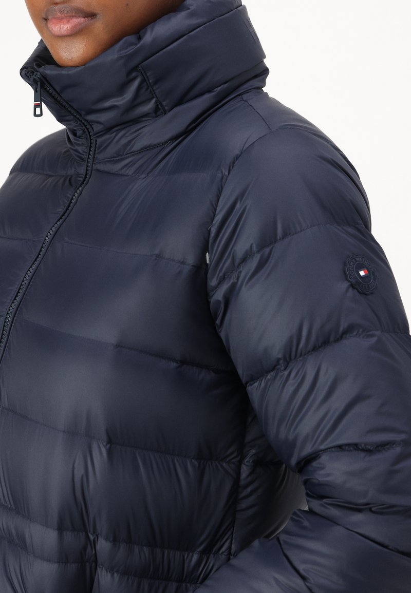 Marineblaue gepolsterte Jacke mit hohem Kragen, Reißverschluss und horizontal gestepptem Muster; verfügt über ein Logopatch am Ärmel.