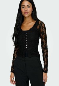Sort blonde cardigan med lange ærmer, der har en bølget halsudskæring og perleknapper foran. Båret over en sort top.