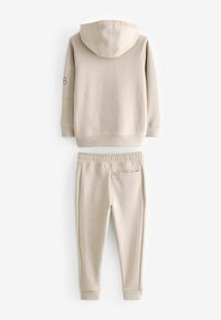 Beige huppari ja siihen sopivat verkkarit, jotka on valmistettu pehmeästä kankaasta. Hupparissa on nyöri ja ribbireunat; verkkareissa on joustava vyötärö ja takatasku.