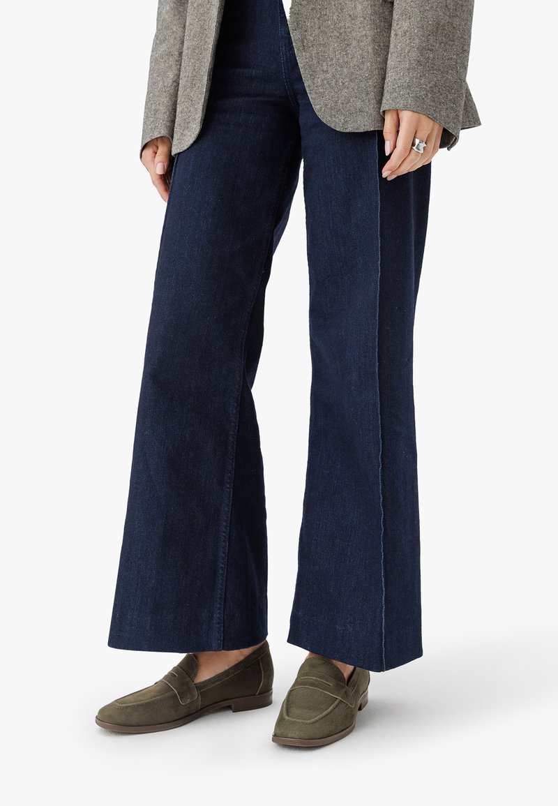 Tumedates sinised laiad püksid, mis on valmistatud denimist, puhta viimistluse ja kontrastsete õmblustega. Koos oliivroheliste nahkkingadega.