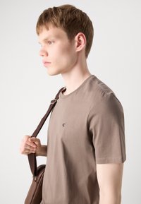 Braunes Baumwoll-T-Shirt mit Rundhalsausschnitt und kurzen Ärmeln, mit einem kleinen schwarzen Logo. Das Model hält eine braune Tasche mit einem Schulterriemen.
