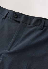 Primo piano di pantaloni formali blu scuro con vita abbottonata e passanti per cintura, che mostrano dettagli di tessuto finemente strutturato.