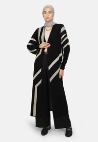 Long black cardigan with beige diagonal stripes, knitted texture, open front, and ribbed cuffs, worn over a light top and loose black pants.