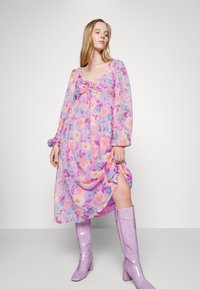 Vestido midi floral em rosa pastel e roxo com mangas longas, decote em forma de barco e detalhe de laço na cintura; apresenta uma fenda lateral.
