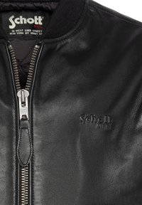 Chaqueta de cuero negro con cremallera plateada, logo en relieve "Schott NYC" en el pecho y etiqueta de tela que muestra la marca y la dirección de Nueva York dentro del cuello.