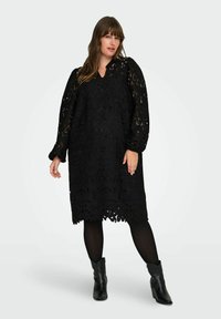 Žena pozira u crnoj čipkastoj midi haljini s balon rukavima, crnim tajicama i crnim čizmama do gležnjeva na jednobojnoj svijetloj pozadini.