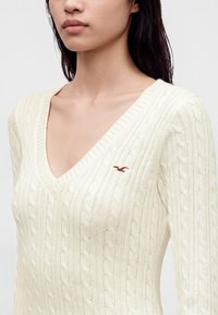 Femme portant un pull à col en V en maille torsadée couleur crème avec un petit logo brodé d’oiseau marron sur la poitrine gauche.