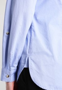 Ljusblå skjorta med långa ärmar, knäppta manschetter och sidsprund. Materialet verkar slätt med en subtil textur.