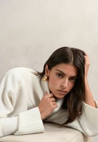 Pletený krémový sveter s vysokým, rebrovaným golierom; v páre s veľkými, lesklými zlatými náušnicami. Ruka modelky spočíva na brade, ukazujúc hladkú pleť.