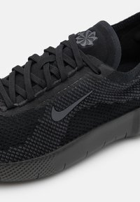 Schwarzer Sportschuh mit einem Obermaterial aus strukturiertem Stoff und flachen Schnürsenkeln. Mit einem schwarzen Nike-Swoosh und einem dezenten Logo-Detail in der Nähe des Kragens.