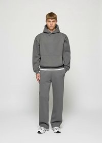 Jauns vīrietis stāv taisni, ģērbies pelēkā kapucē un sporta biksēs, ar baltām kedām, uz viendabīga gaiša fona.