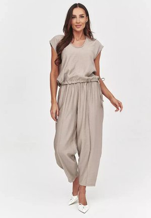 Kobieta nosząca luźną beżową bluzkę z krótkimi rękawami typu czapka i szerokie spodnie, w połączeniu z białymi szpiczastymi szpilkami, stojąca na jednolitym tle.