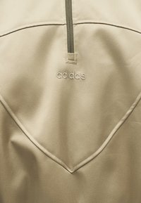 Κοντινή λήψη ενός μπεζ ενδύματος Adidas με κοντό κάθετο φερμουάρ και κεντημένο λογότυπο Adidas κάτω από το φερμουάρ.