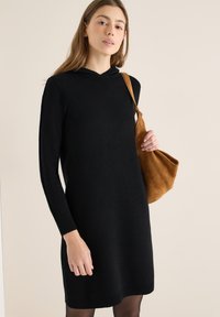 Vestito nero in maglia con cappuccio, maniche lunghe e polsini a coste. Indossato con una borsa a tracolla in suede marrone. Tessuto liscio, design casual.