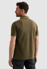 Hombre de perfil hacia la izquierda, vistiendo una camisa polo de manga corta color verde oliva y jeans oscuros, sobre un fondo liso gris claro.