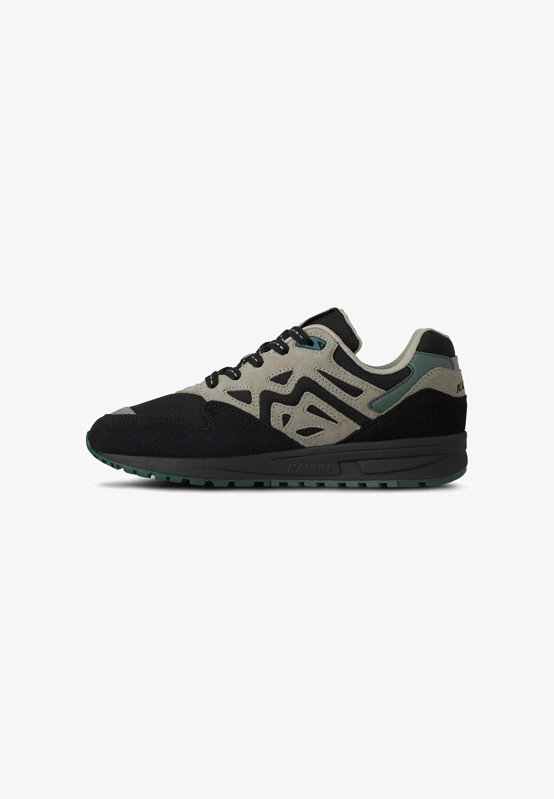 Sneaker bassa nera e grigia con sovrapposizioni in suede con motivo, lacci neri e suola scura con texture su sfondo bianco.