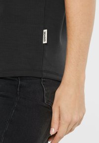 Right hand resting by side, wearing black shirt with "SENSES" tag and black jeans against plain background.