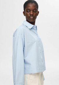 Camicetta blu chiara con colletto, vestibilità oversize e maniche lunghe. Presenta una tasca sul petto e un tessuto liscio e testurizzato.