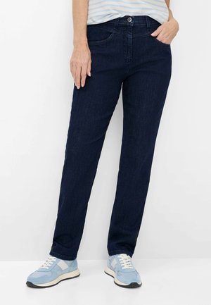 Frau trägt dunkelblaue Jeans mit geradem Beinschnitt, hellblau-weiße Turnschuhe und ein in die Hose gestecktes gestreiftes Oberteil, steht vor weißem Hintergrund.