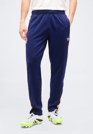 Persona vistiendo pantalones deportivos Adidas azul marino con rayas amarillas, zapatos de tacos blancos con suelas verde neón, de pie contra un fondo liso.