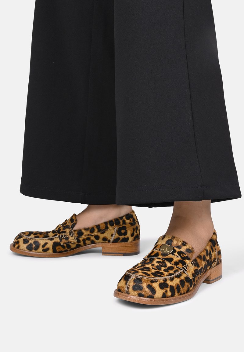 Mocassini con stampa leopardata, texture liscia, tacco in legno marrone e un piccolo dettaglio metallico. Indossati con pantaloni neri a gamba larga.