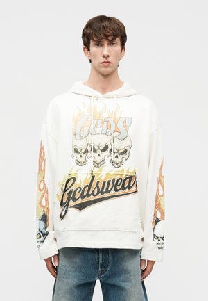 Młody mężczyzna ubrany w białą bluzę z kapturem z grafiką płonących czaszek i napisem "Gcdswear", w połączeniu z niebieskimi dżinsami, stoi na jednolitym tle.